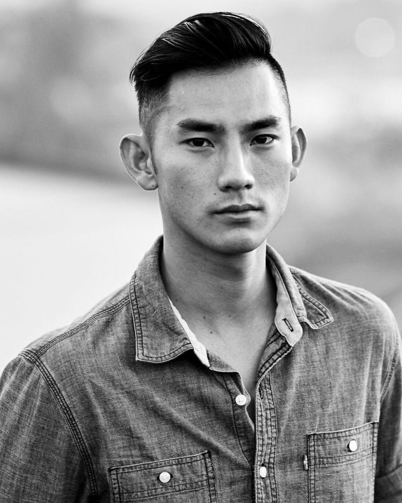 Black and white portrait of a young man with short, styled hair, wearing a denim jacket, outdoors with a blurred background.
