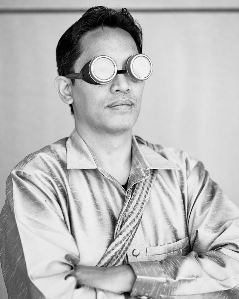 Black and white photo of a man with arms crossed, wearing goggles and a shiny shirt, with a serious expression.