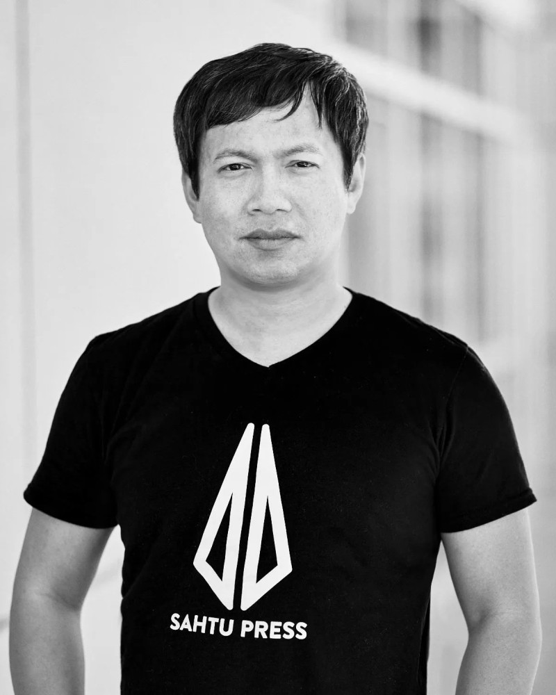 Black and white photo of a man wearing a black T-shirt with Sahtu Press logo, standing indoors with blurred background.