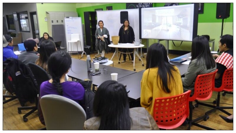 Two women present at the front of a classroom to seated participants during a workshop