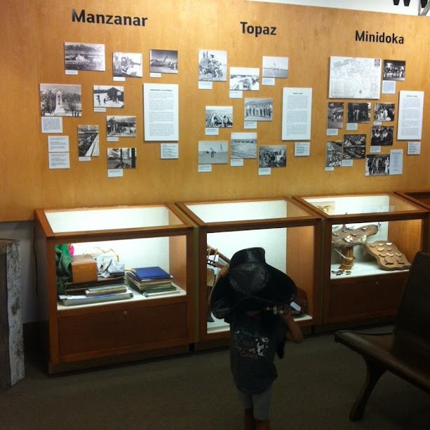 Museum display on Manzanar, Topaz, and Minidoka incarceration camps, with a child in a wide hat in front