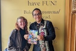 Two people smiling at a bakery, one holding up a book