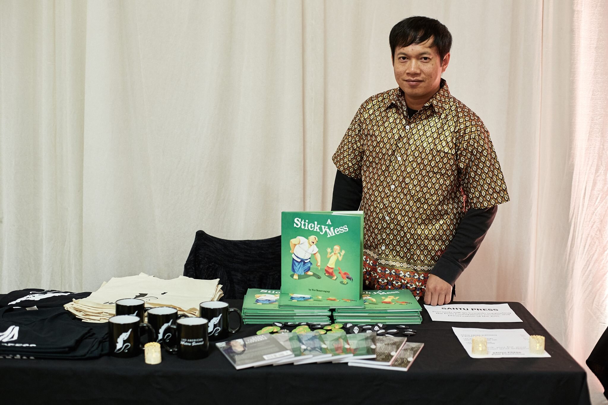 Nor Sanavongsay standing behind a Sahtu Press table with copies of A Sticky Mess, mugs, and T-shirts.