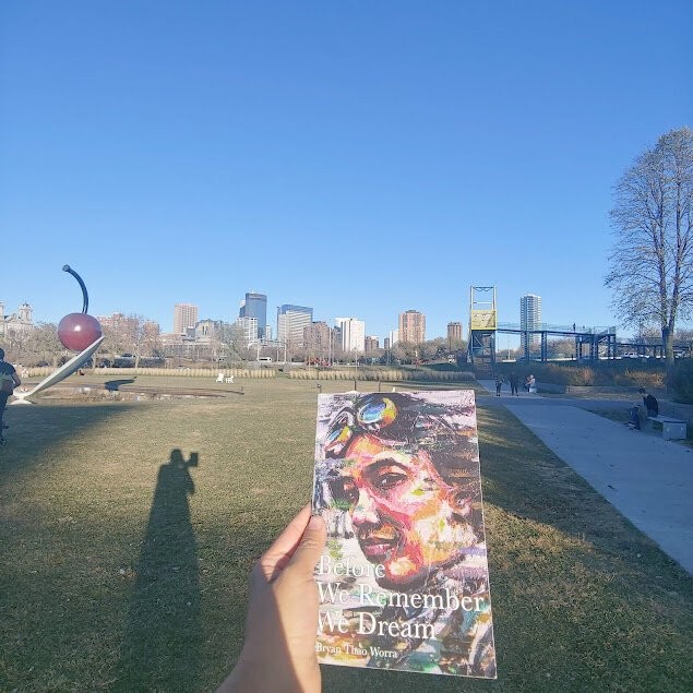 Hand holding Before We Remember We Dream by Bryan Thao Worra, its cover a colorful painted face, at the Minneapolis Sculpture Garden.