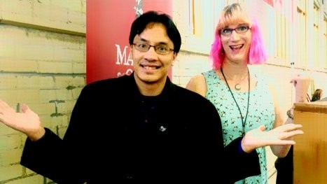Bryan Thao Worra and Charlie Jane Anders smiling and shrugging playfully in front of a red banner.
