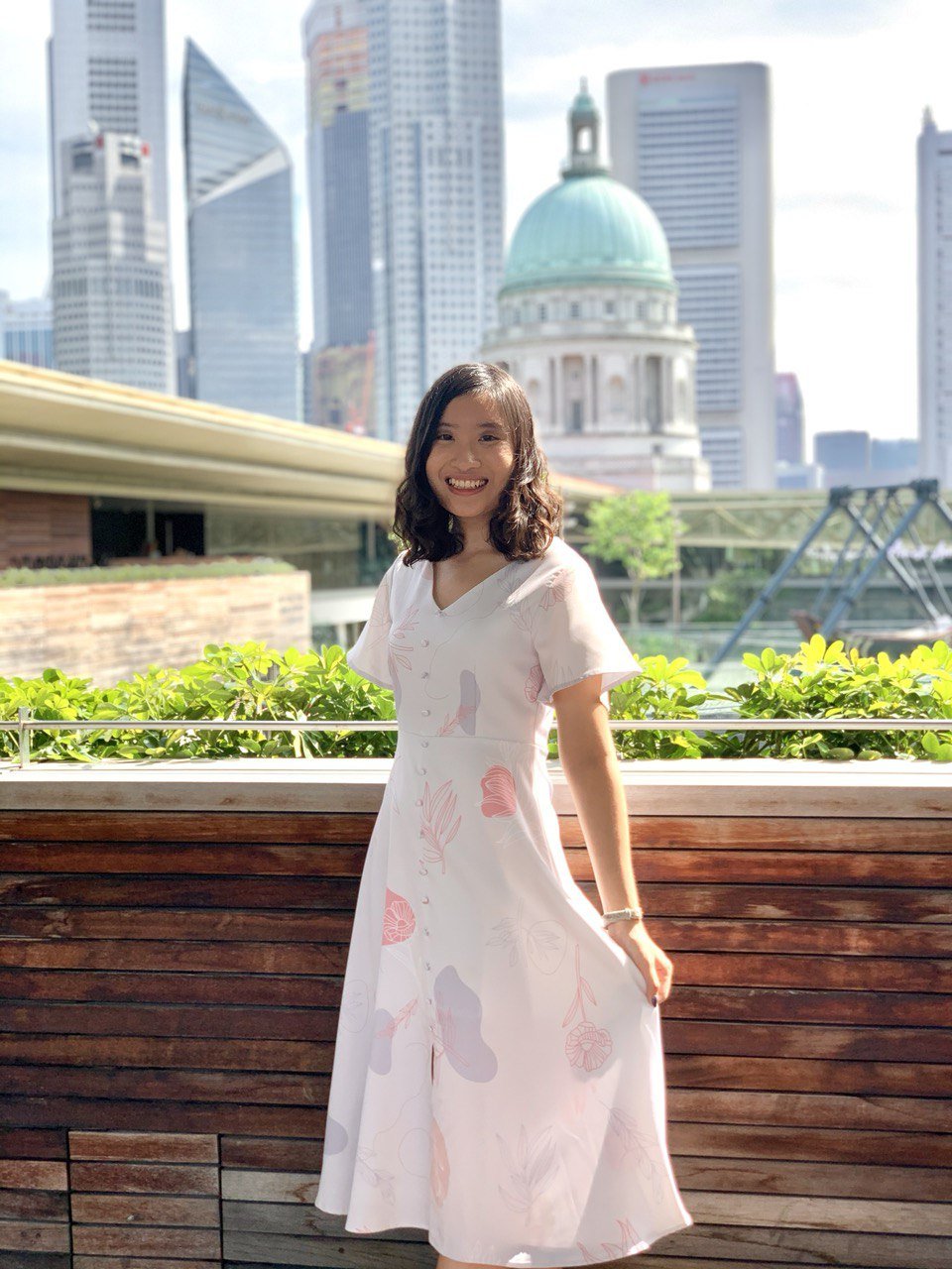 Sarah Ang in a white floral dress on a terrace, with Singapore skyscrapers and a green dome behind her