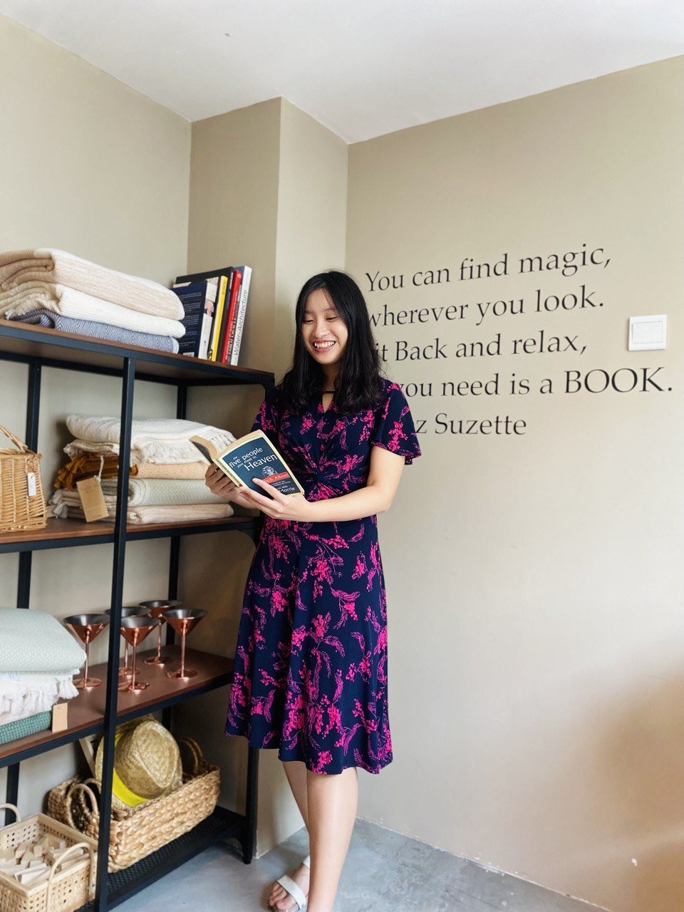 Sarah Ang smiling as she reads a book beside shelves, under a wall quote about finding magic in a book