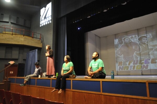 Four presenters seated and standing on the edge of a UMass Lowell stage, with 'Refugee Nation' projected behind them.
