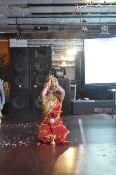 Dancer in gold and red traditional costume and headdress kneeling on a stage with hands raised together above her head.