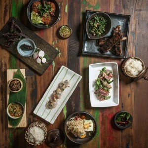 Overhead spread of Lao dishes from Khe-Yo on a painted wood table, including grilled meats, greens, dipping sauces, and sticky rice