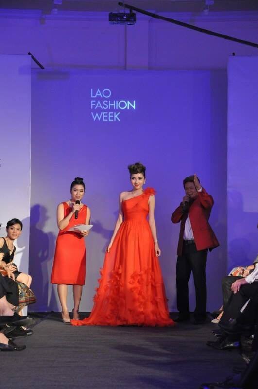 Model in a flowing red gown on stage at Lao Fashion Week, between a woman with a microphone and a man in a red jacket