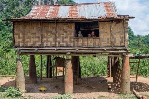 Family looking out the window of a bamboo-walled stilt house with a tin roof in rural Laos