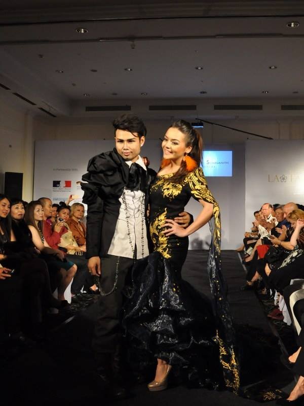 Male and female models on a fashion runway in black and gold embroidered outfits, audience seated alongside