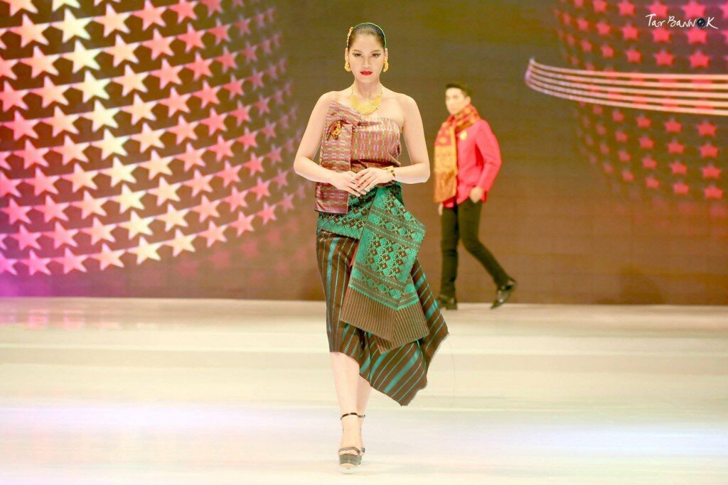 Model on a runway in a Lao silk sinh skirt and woven green shawl, a man in a red jacket behind her
