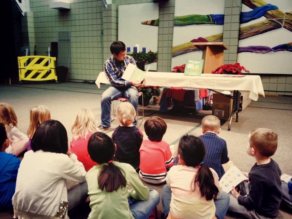 A man reads a picture book aloud to a group of young children seated on the floor