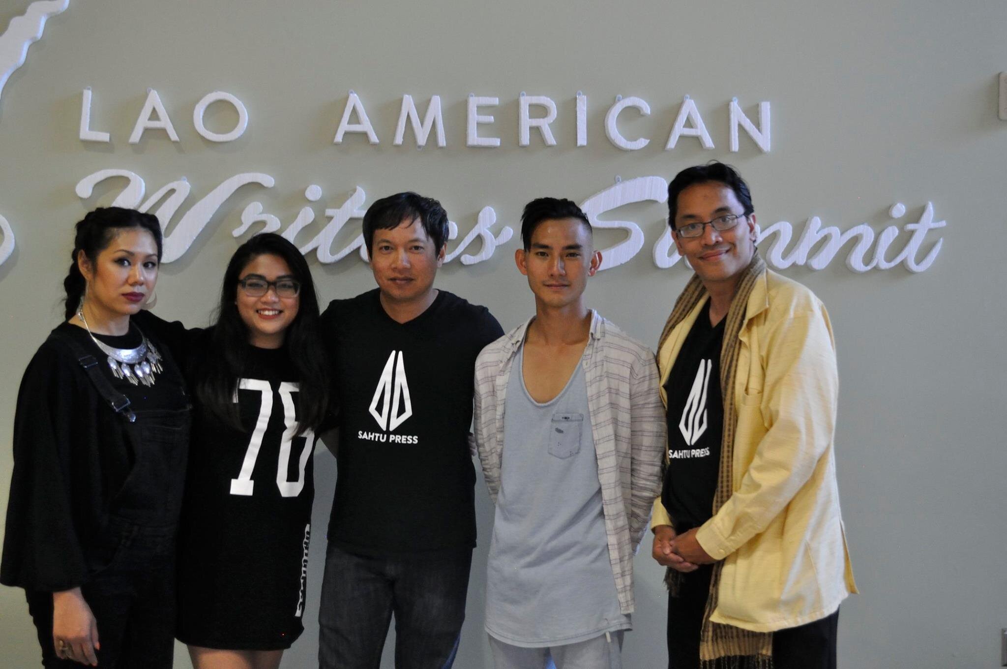 Sahtu Press team, three in black Sahtu Press shirts, posing in front of the Lao American Writers Summit sign