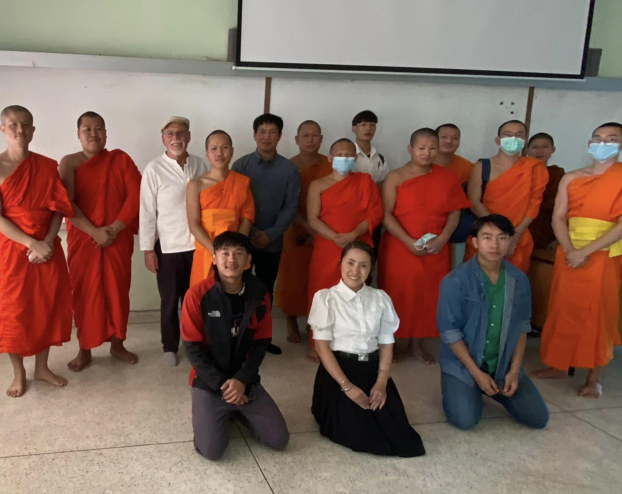 Group photo of Buddhist monks in orange robes with students and visitors in a classroom at Wat Suan Dok