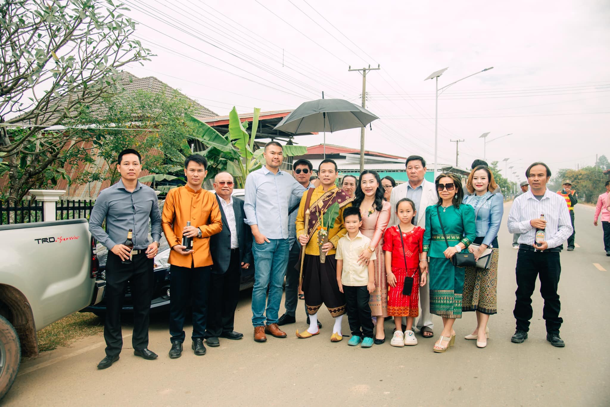 Wedding party in Laos posing on a street, the groom in traditional Lao dress holding a sword, guests in sinh skirts