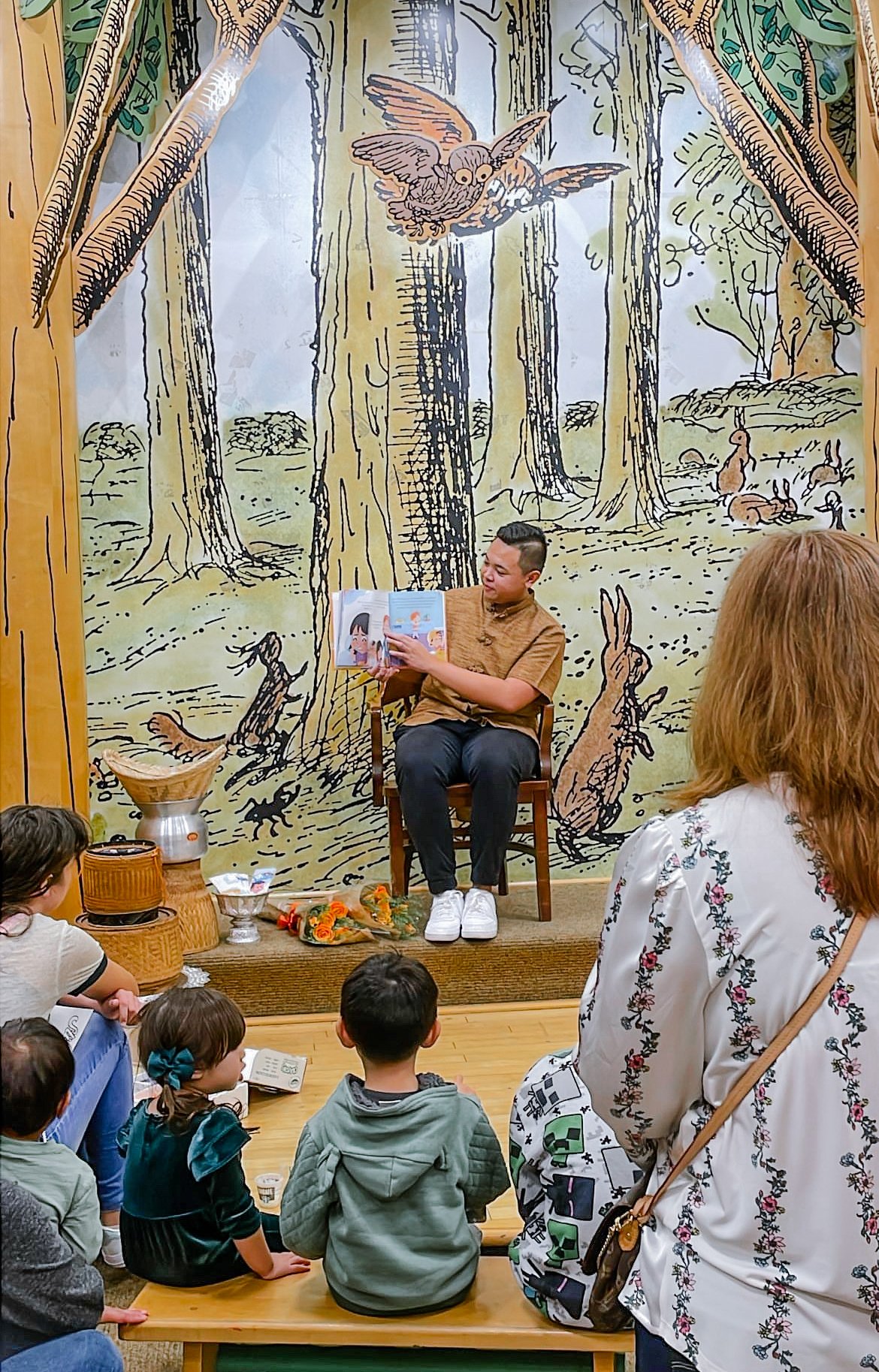 Jacob Singhavong reading Sticky Rice for Lunch to children in front of a forest mural