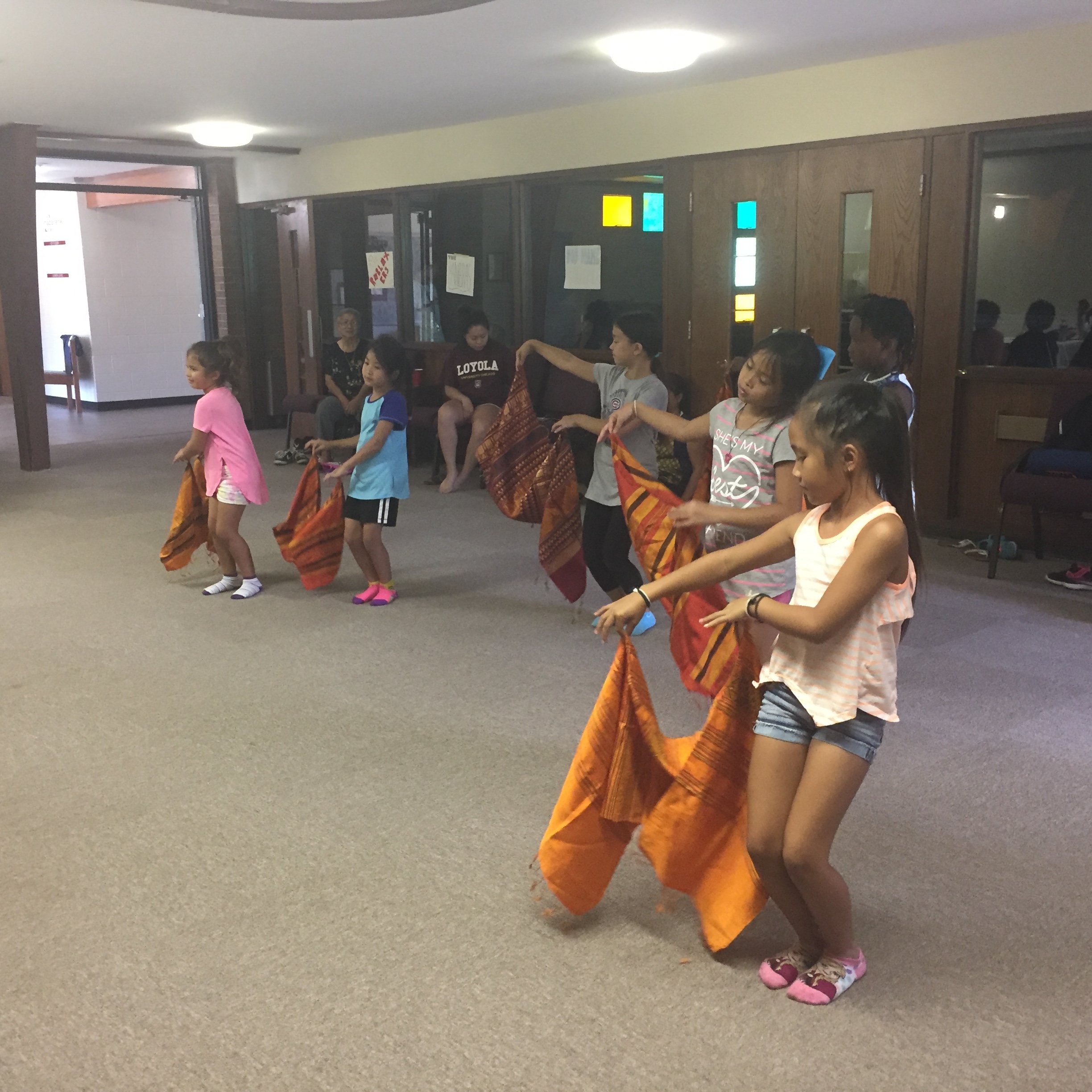 Young girls practicing a Lao dance, each holding an orange woven scarf, as adults watch.