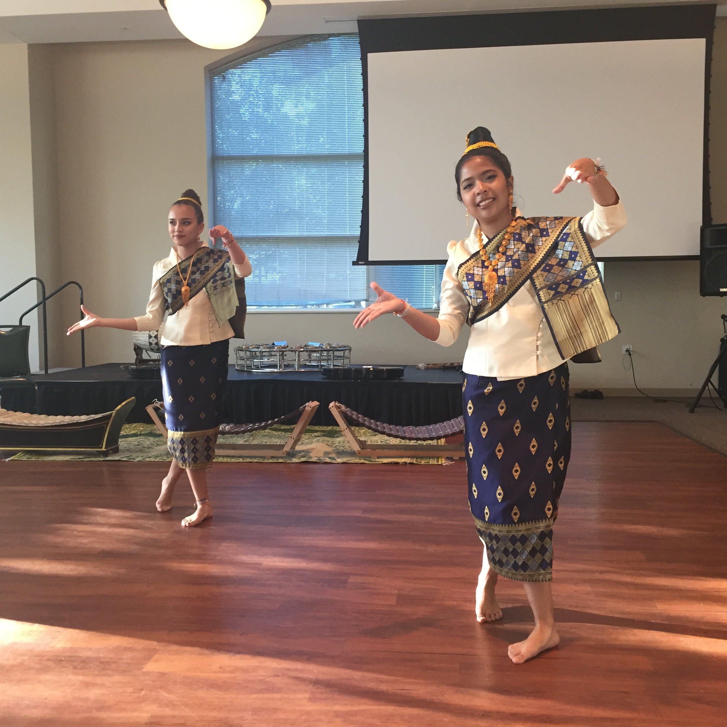 Two young women in blue Lao sinh and white tops performing a traditional Lao dance with graceful hand gestures.