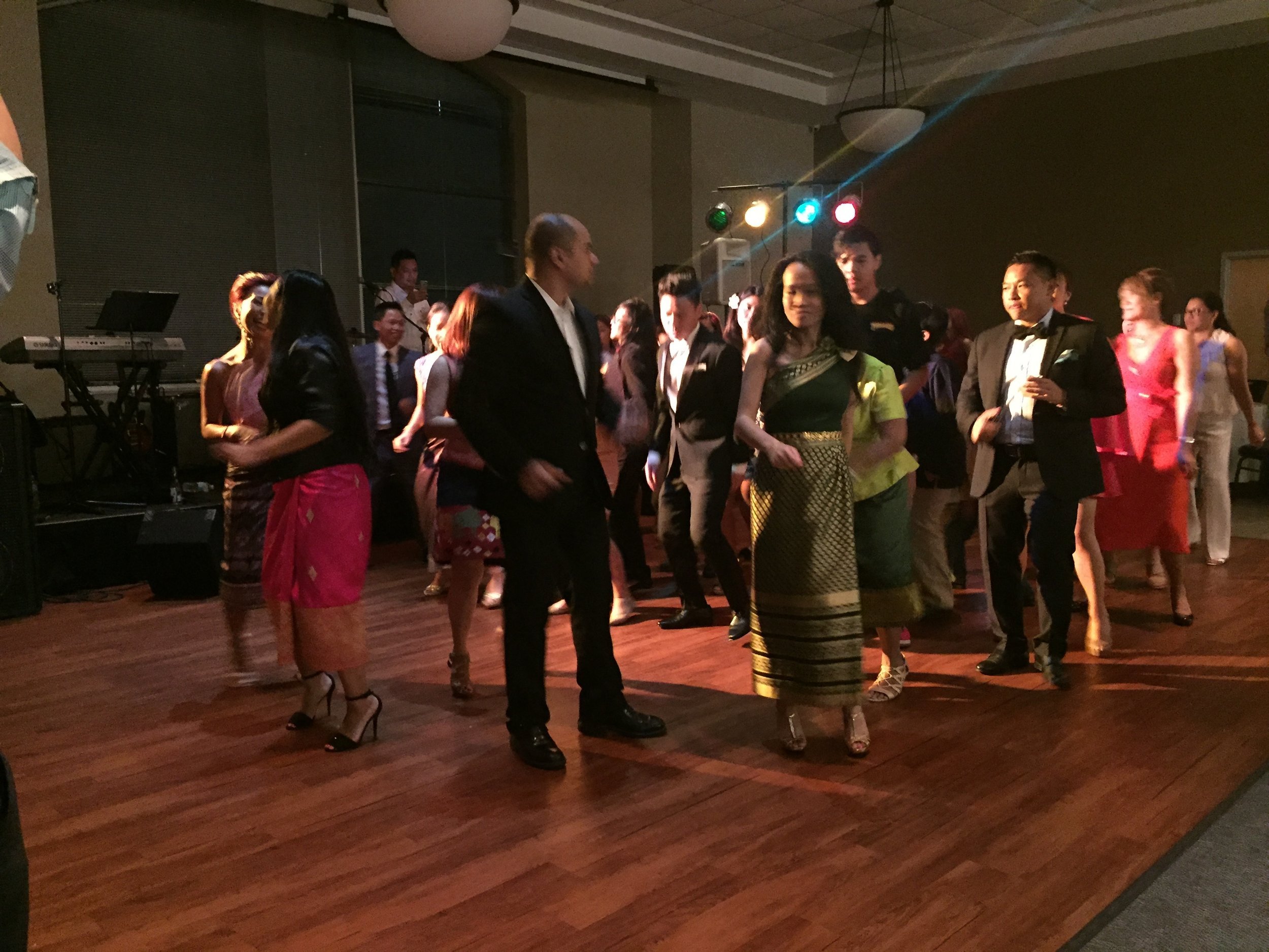 Guests in formal wear and Lao sinh dancing a lamvong line dance in a ballroom.