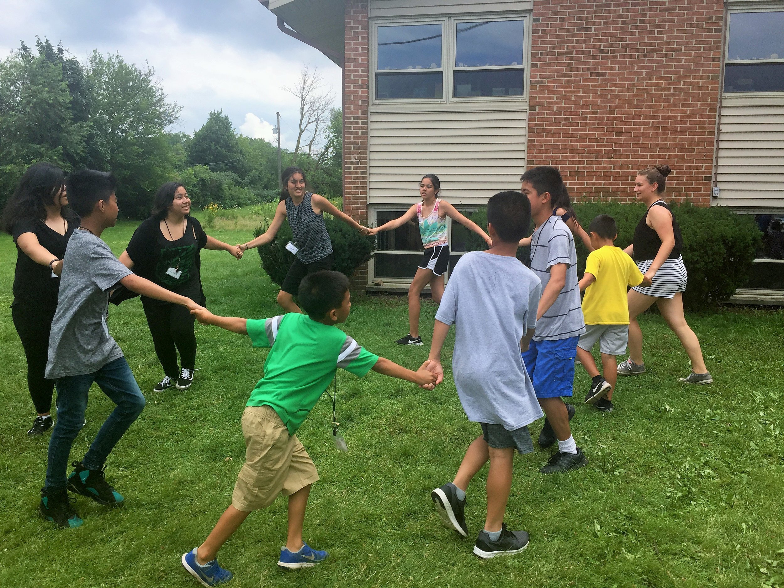 Kids and teens holding hands in a circle on a lawn outside a brick building.