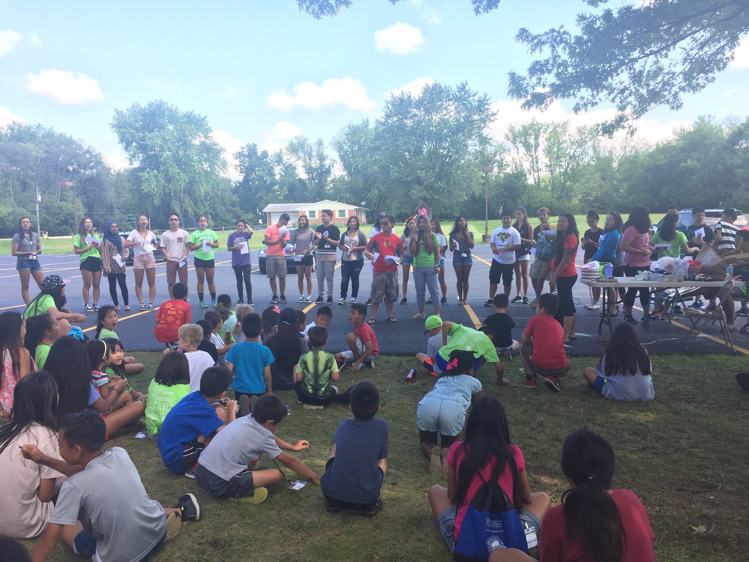 Large group of Lao youth and adults gathered outdoors on grass and pavement for a camp activity.
