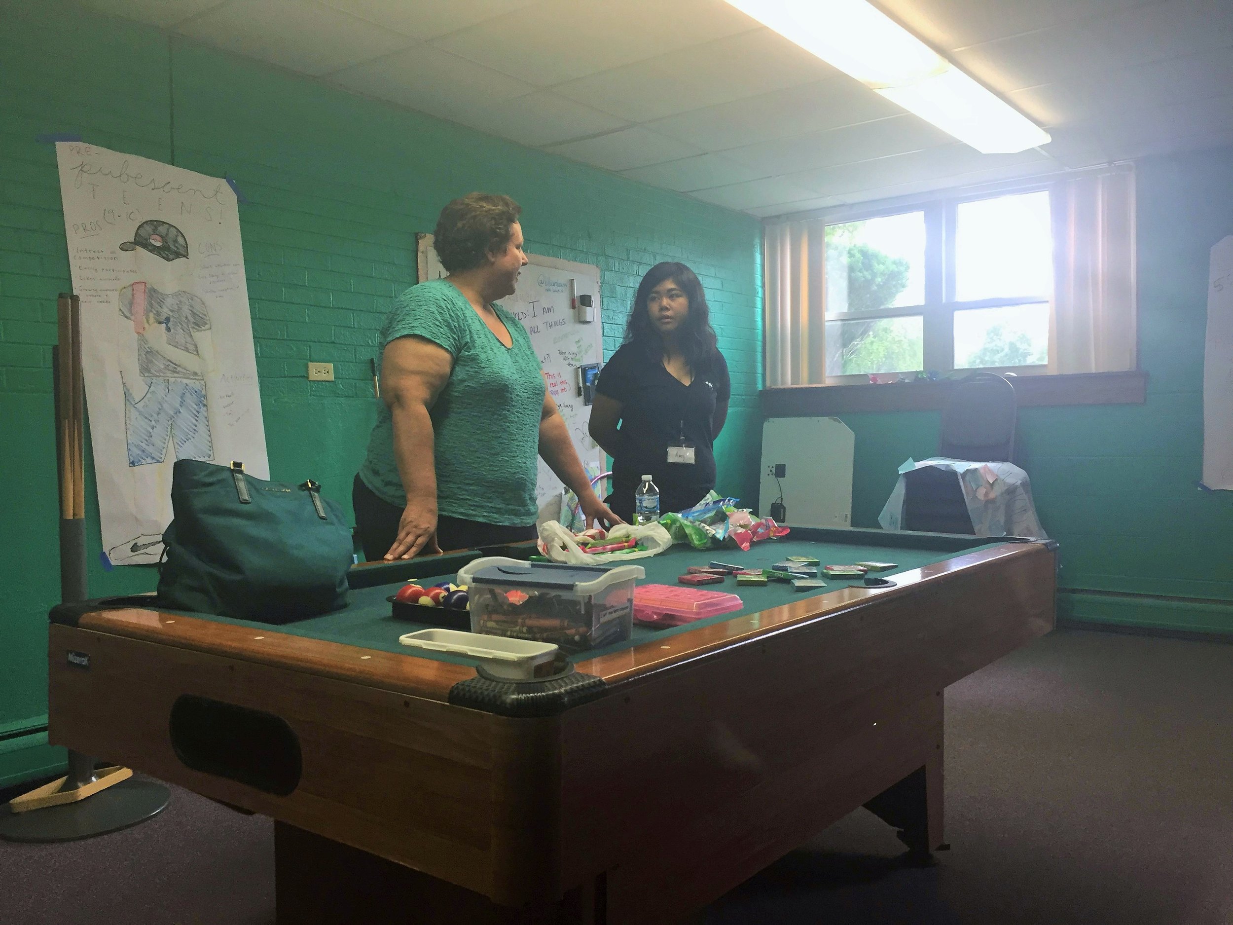Two women talking beside a pool table laid out with craft supplies in a green-walled room.