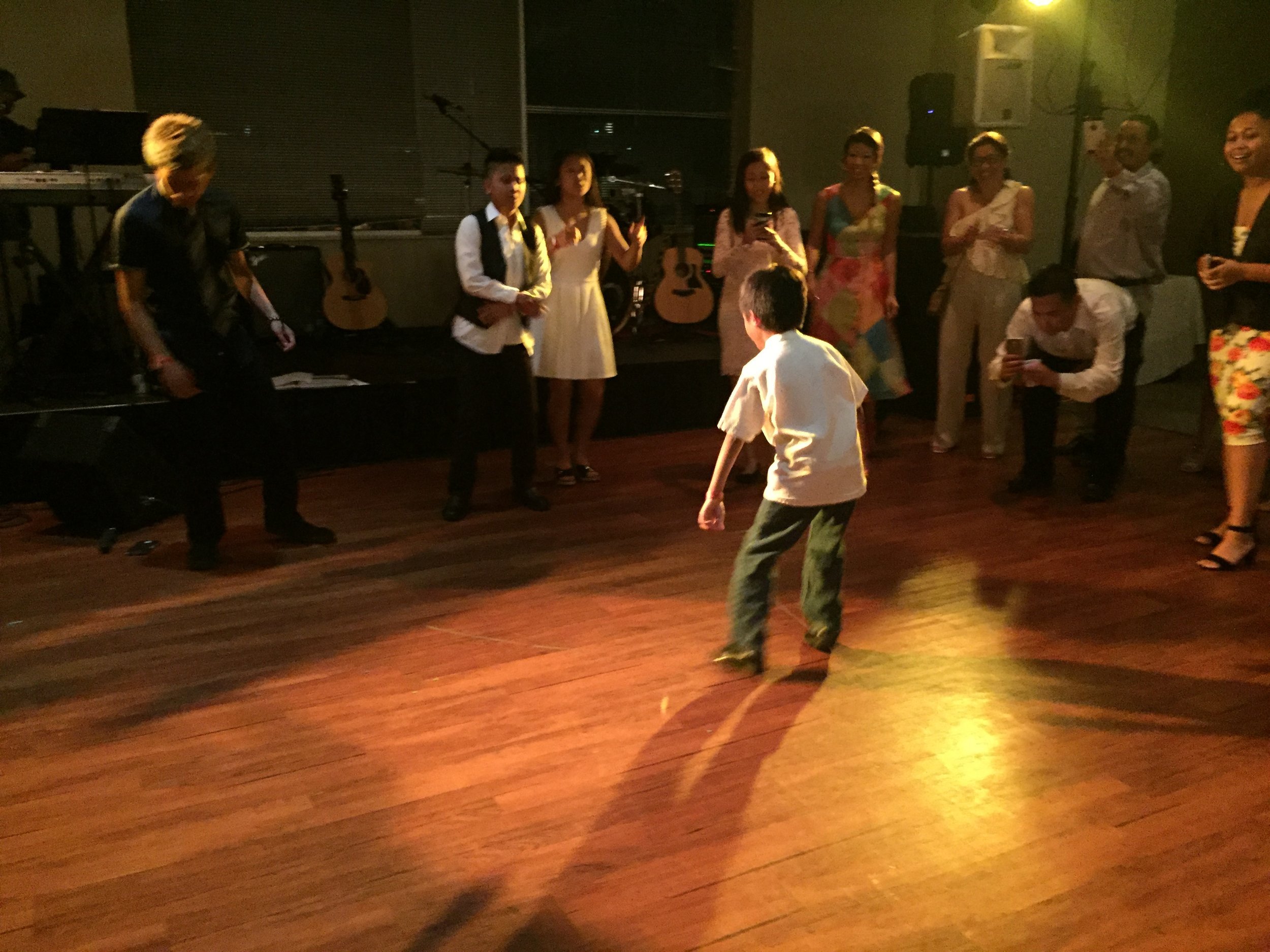 Young boy dancing alone in a spotlight on a wooden floor as guests watch and film with phones.