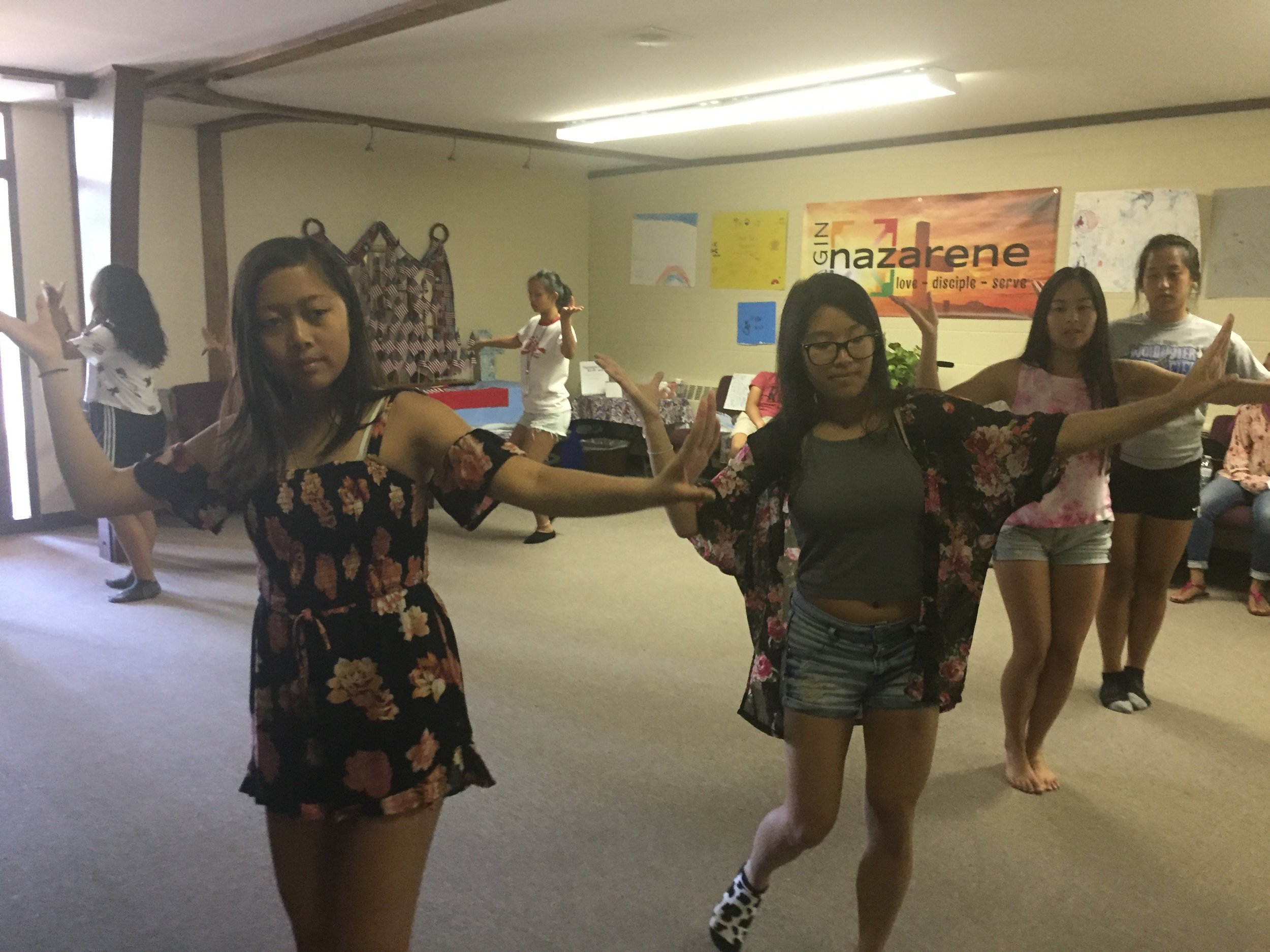 Teen girls practicing a traditional Lao dance with curved hand gestures in a classroom.