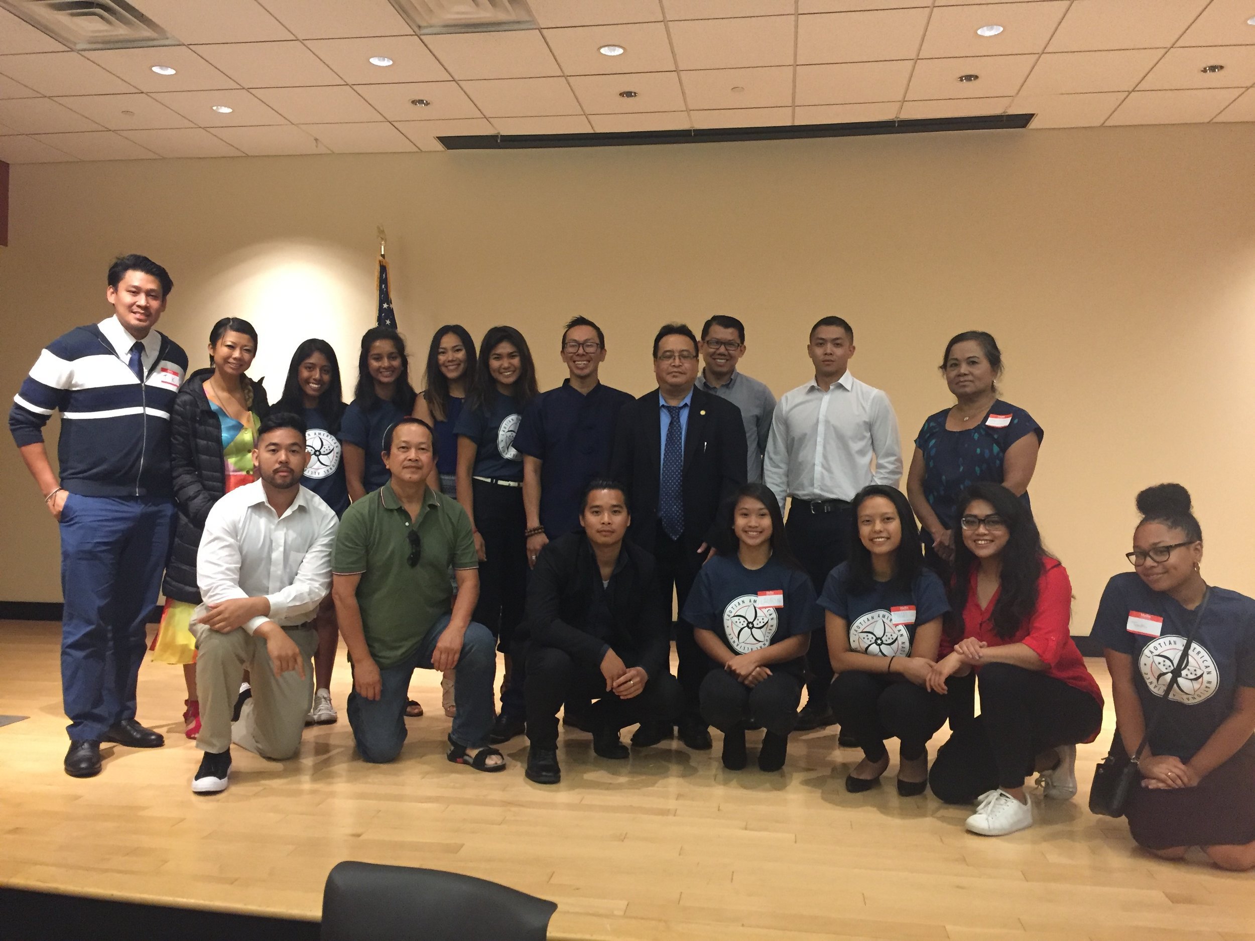 Group photo of about twenty Lao American youth and adults in a meeting room, some kneeling in front.