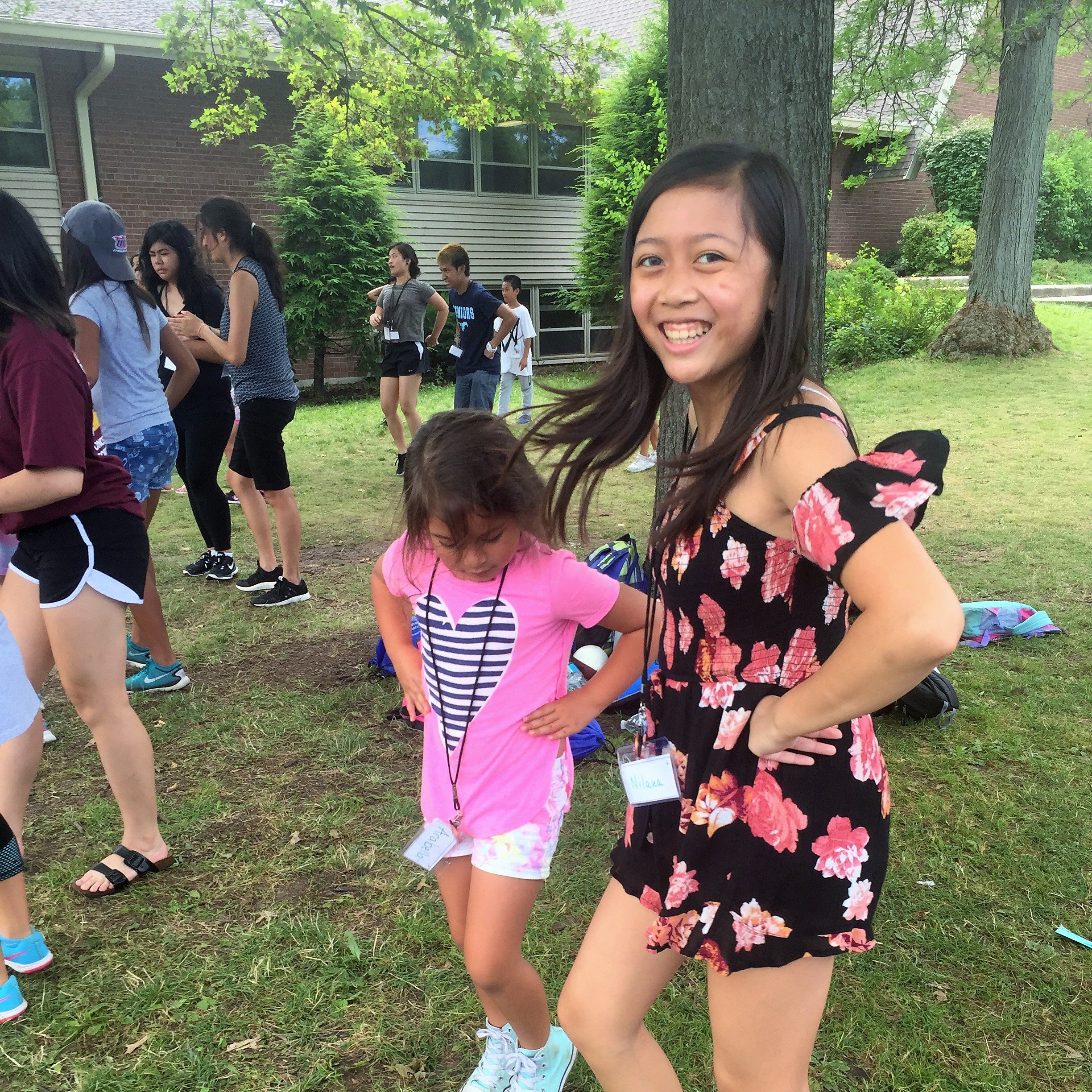 Smiling teen girl in a floral romper posing beside a little girl in a pink shirt at an outdoor camp.