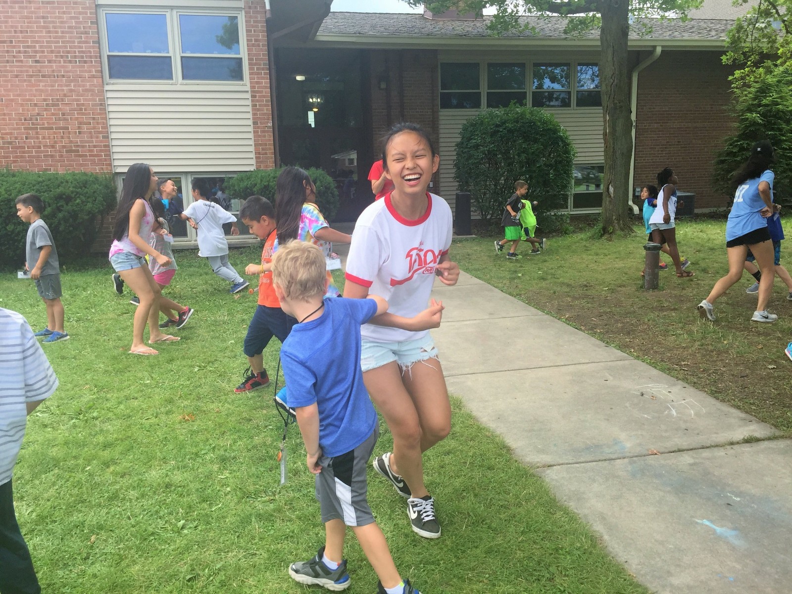 Laughing teen girl in a white T-shirt playing a running game with a young boy on a lawn.