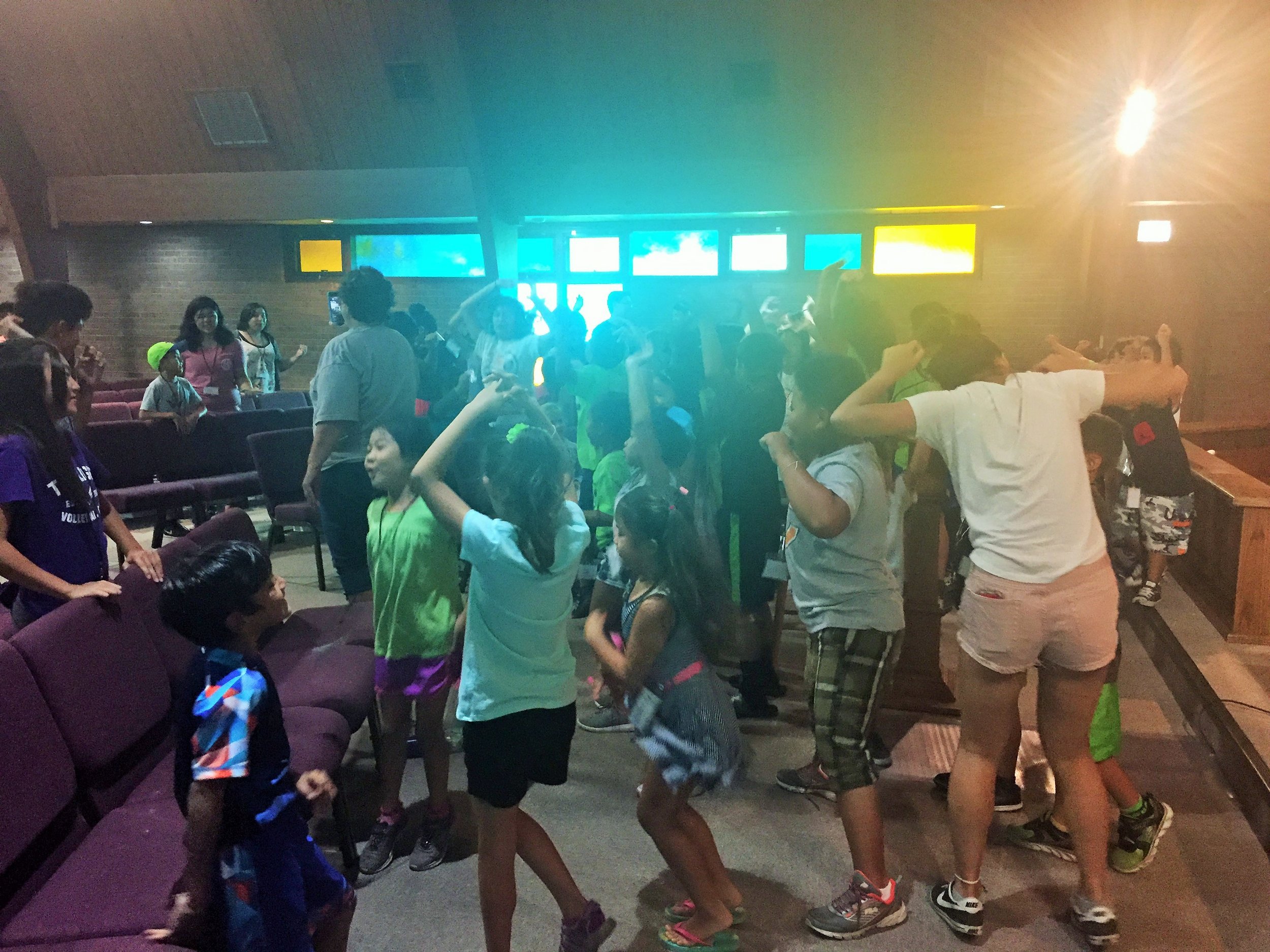 Kids and teens dancing together in a dim hall lit with colorful stage lights.