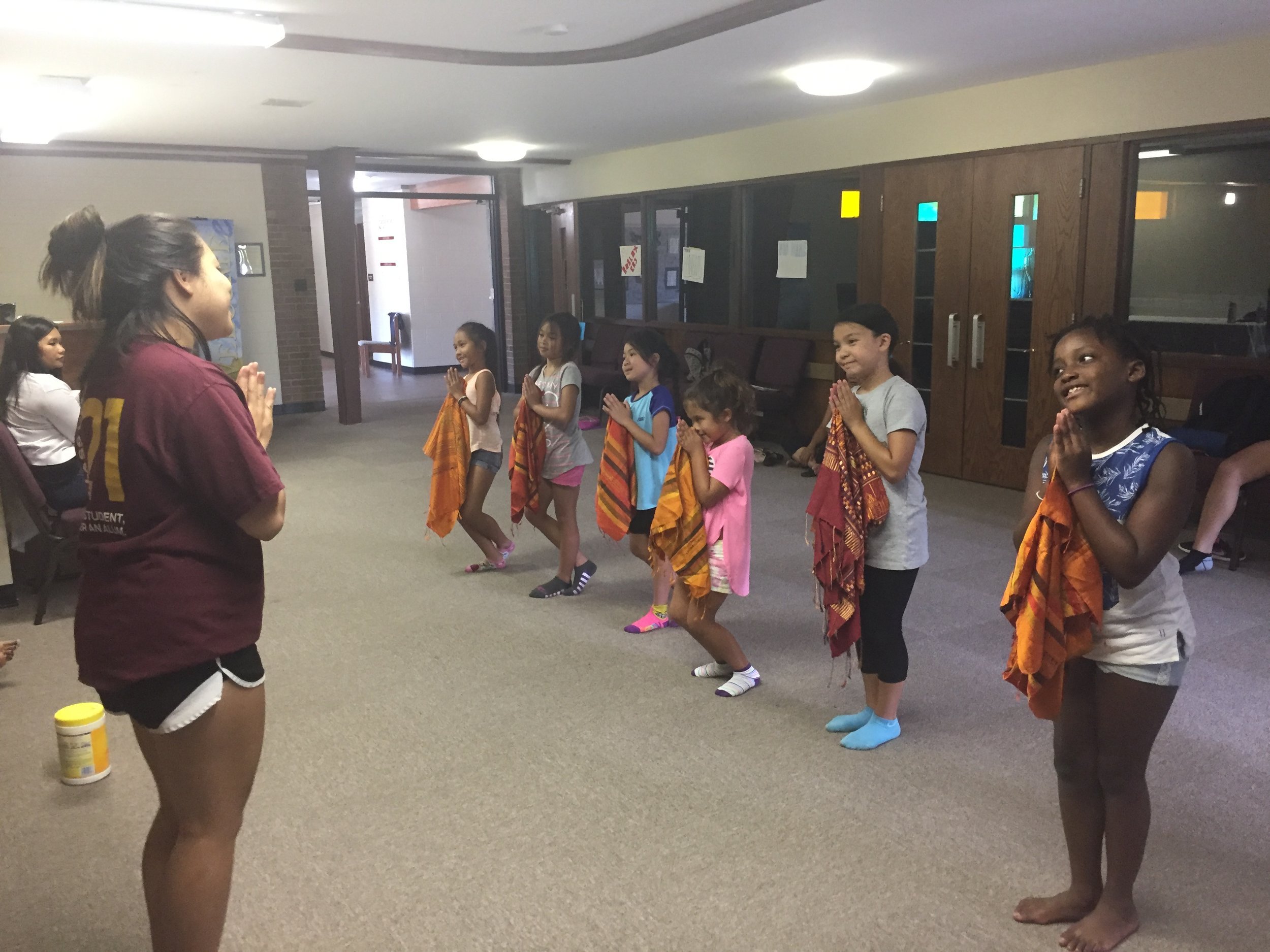 Young woman leading a row of girls holding orange scarves, their hands pressed together in a nop greeting.