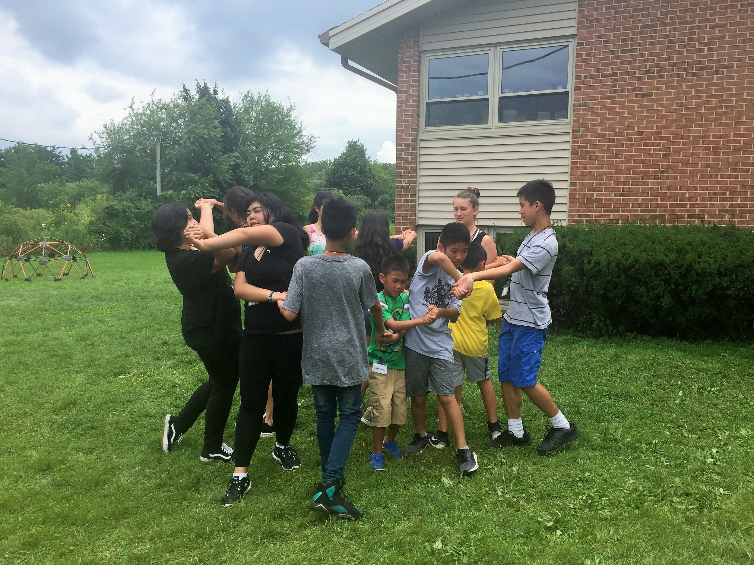 Group of kids and teens tangled together holding hands in a human knot game on a lawn.