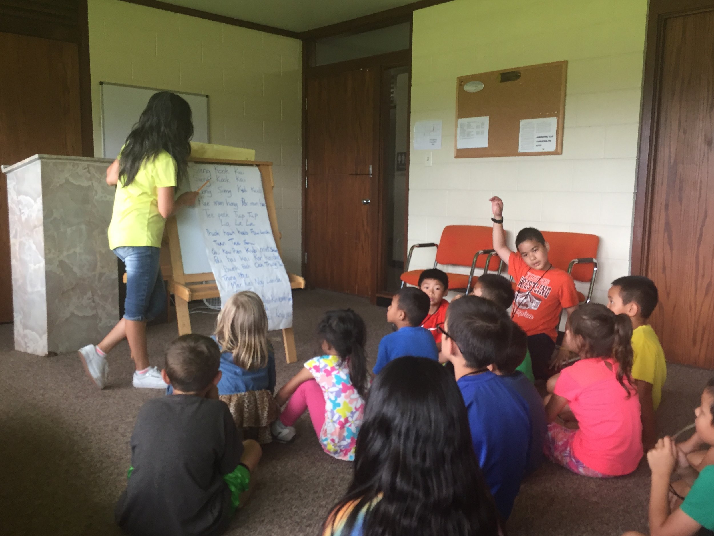 Girl in a yellow shirt writing on an easel pad while seated kids watch and one boy raises his hand.