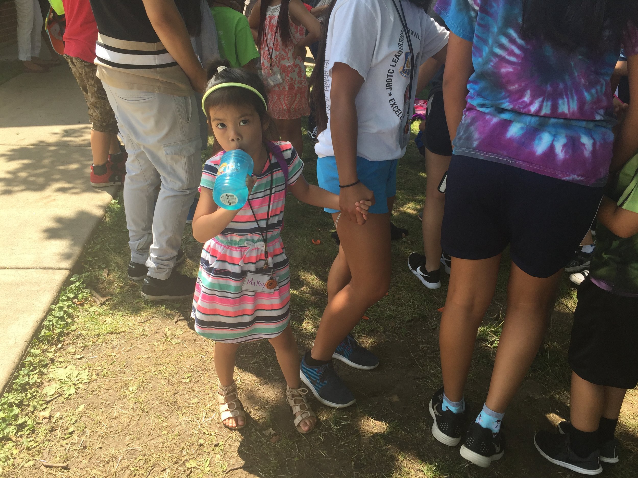 Little girl in a striped dress and headband drinking from a blue cup among older kids outdoors.