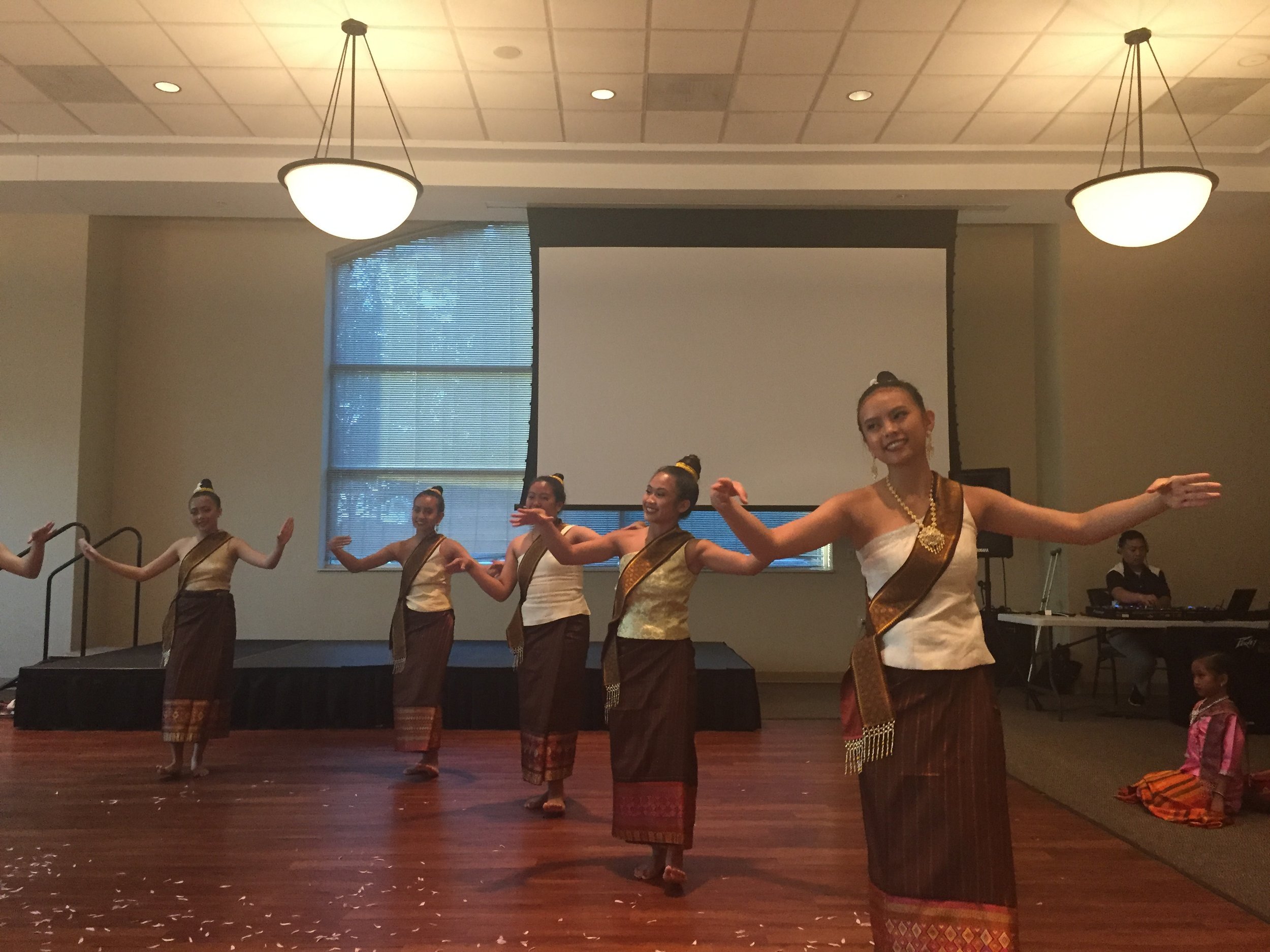 Five young women in Lao sinh and sashes performing a traditional Lao dance with arms outstretched.