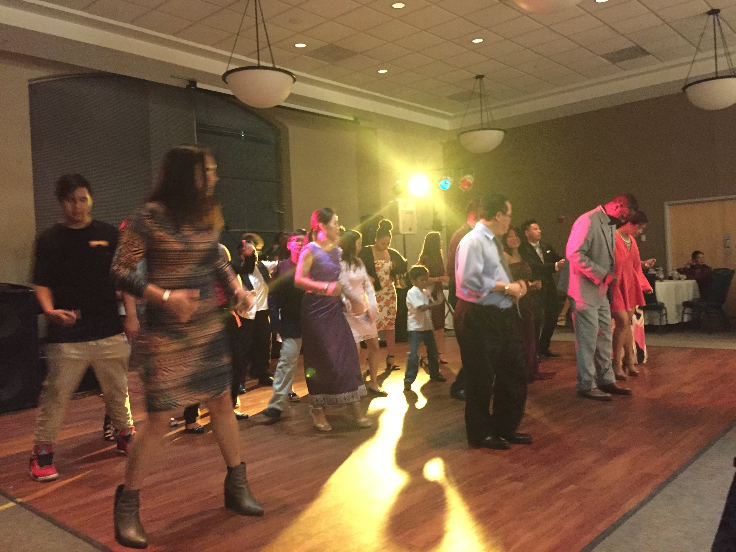 Guests of all ages dancing a lamvong line dance on a ballroom floor under colored lights.