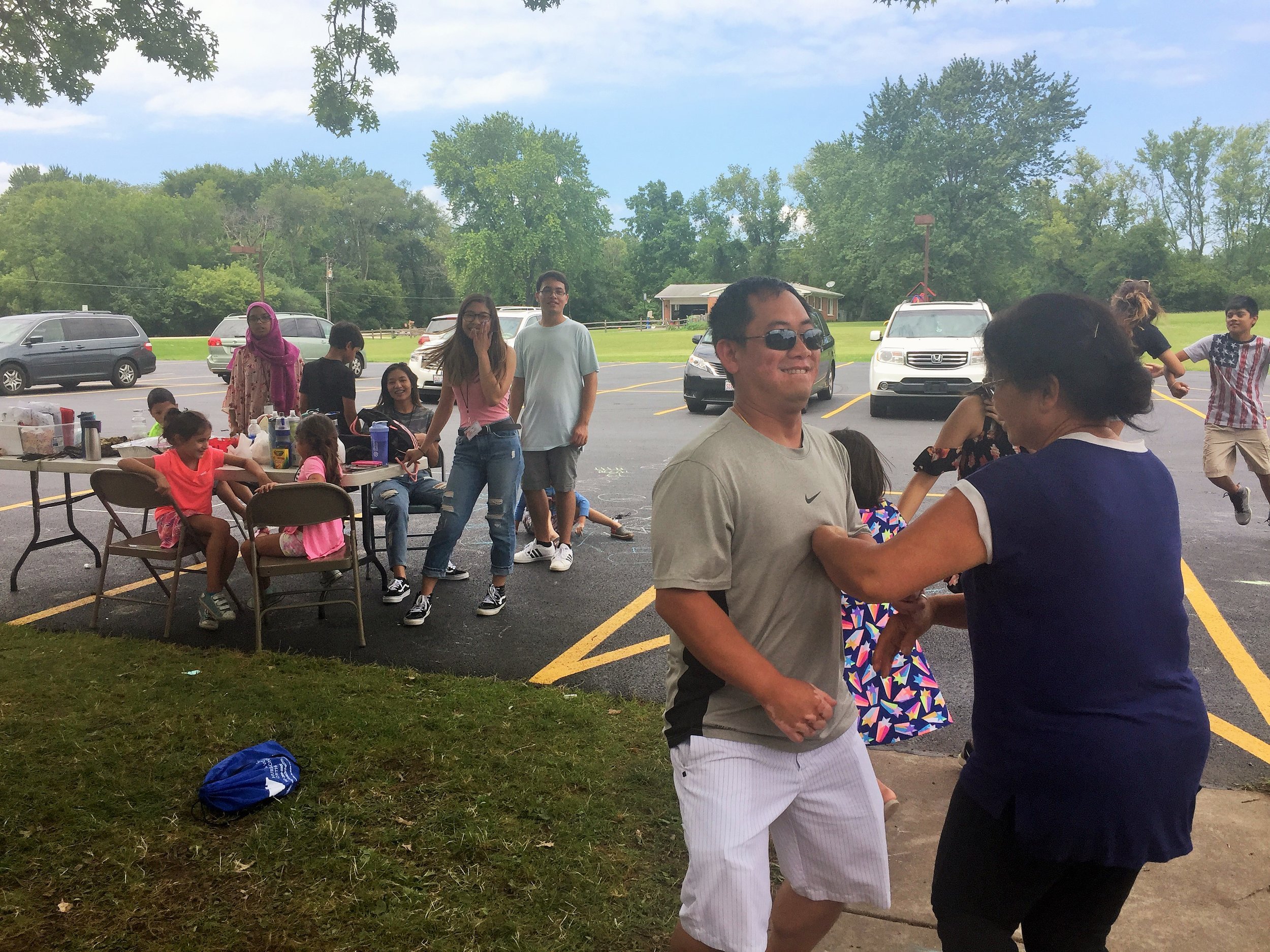 A man and an older woman dancing together in a parking lot at a picnic while others sit at tables.
