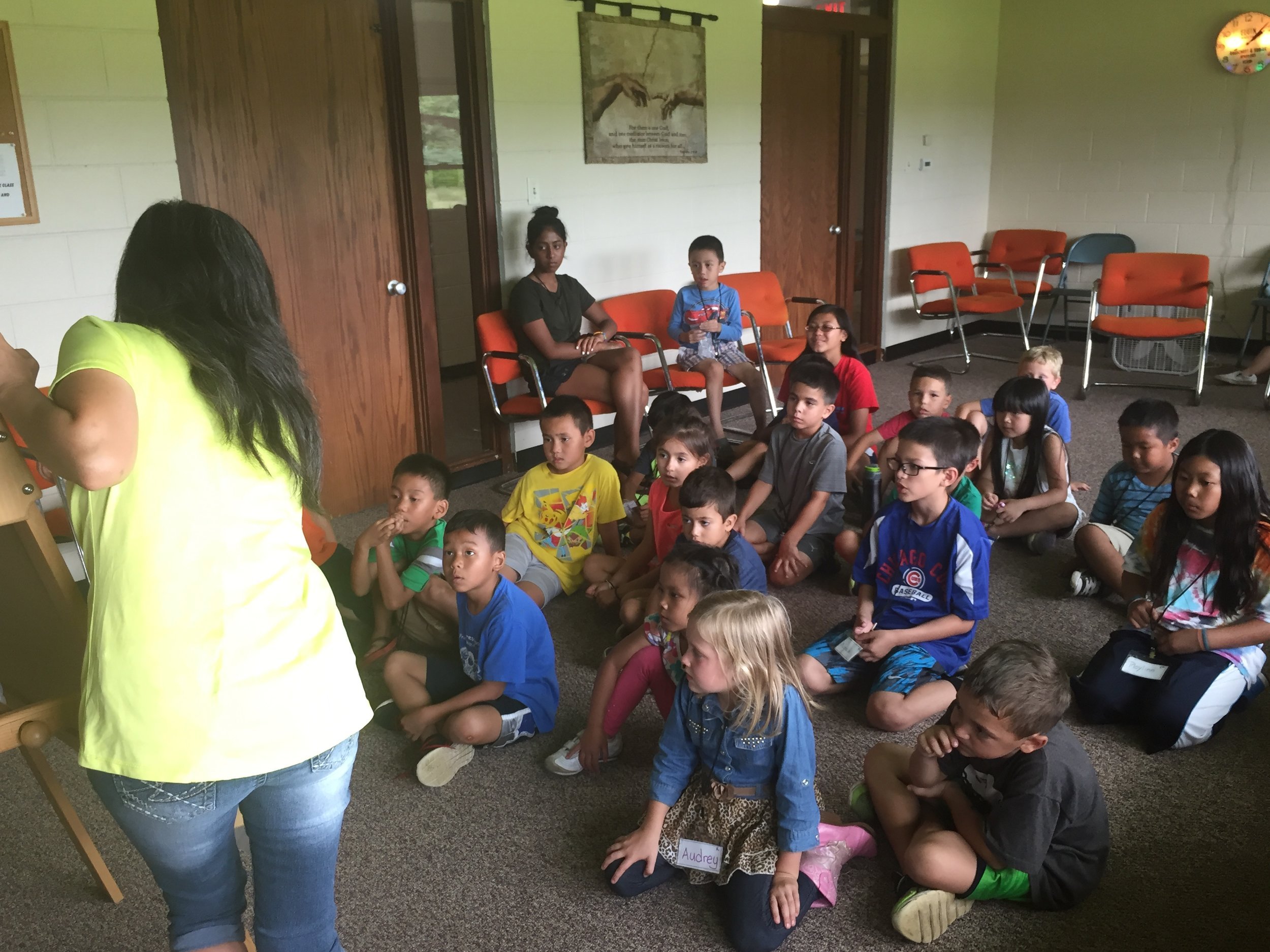 Kids sitting on a carpeted floor watching a woman in a yellow shirt write on an easel pad.
