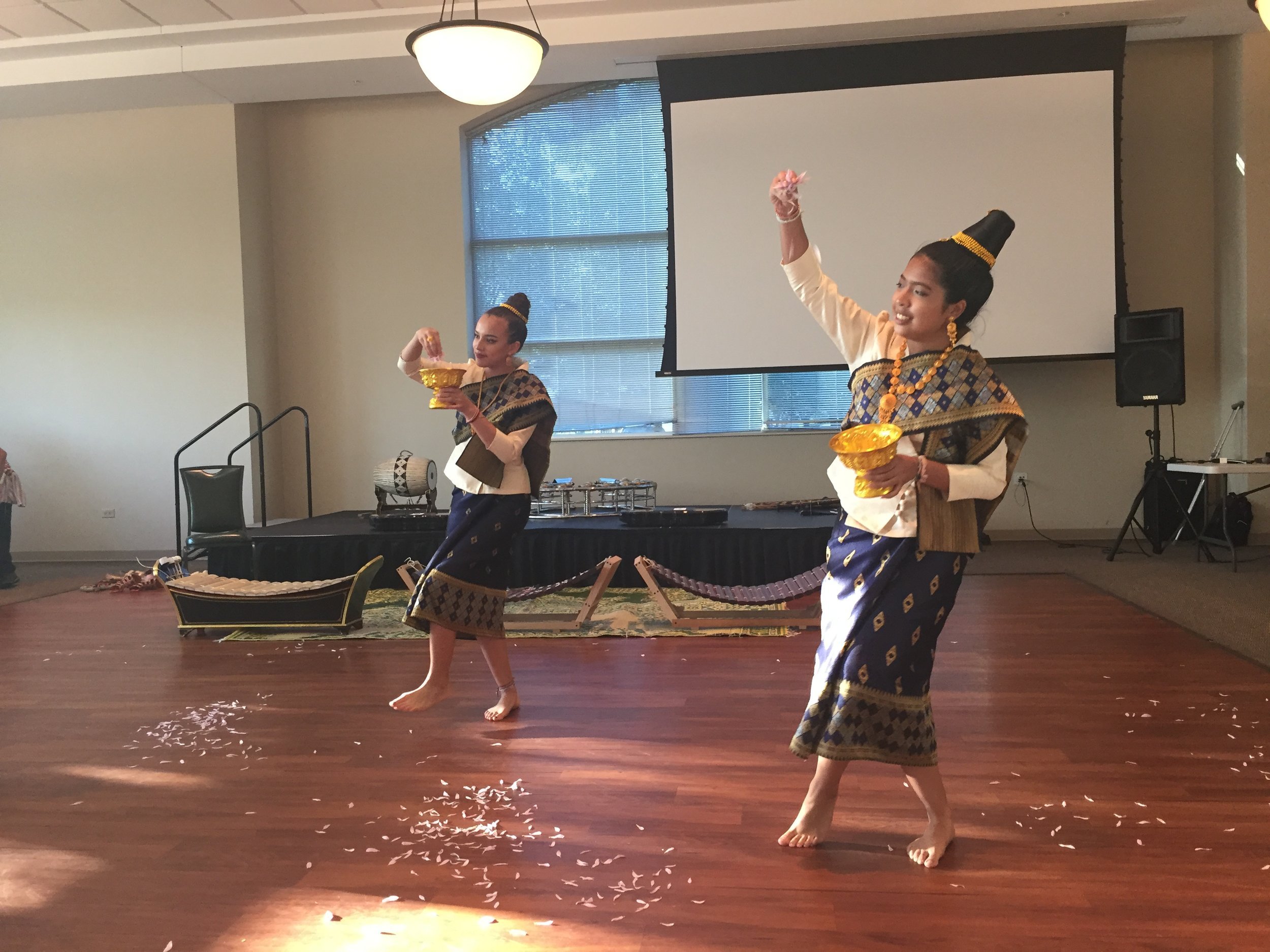 Two dancers in Lao sinh and sashes performing a traditional dance holding gold bowls of flowers.