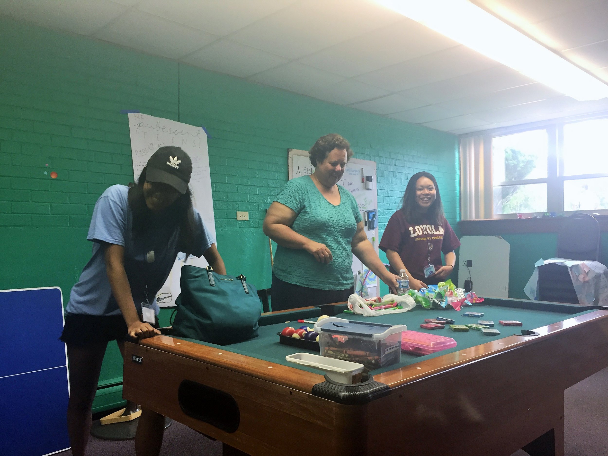 Three women laughing around a pool table covered with craft supplies.