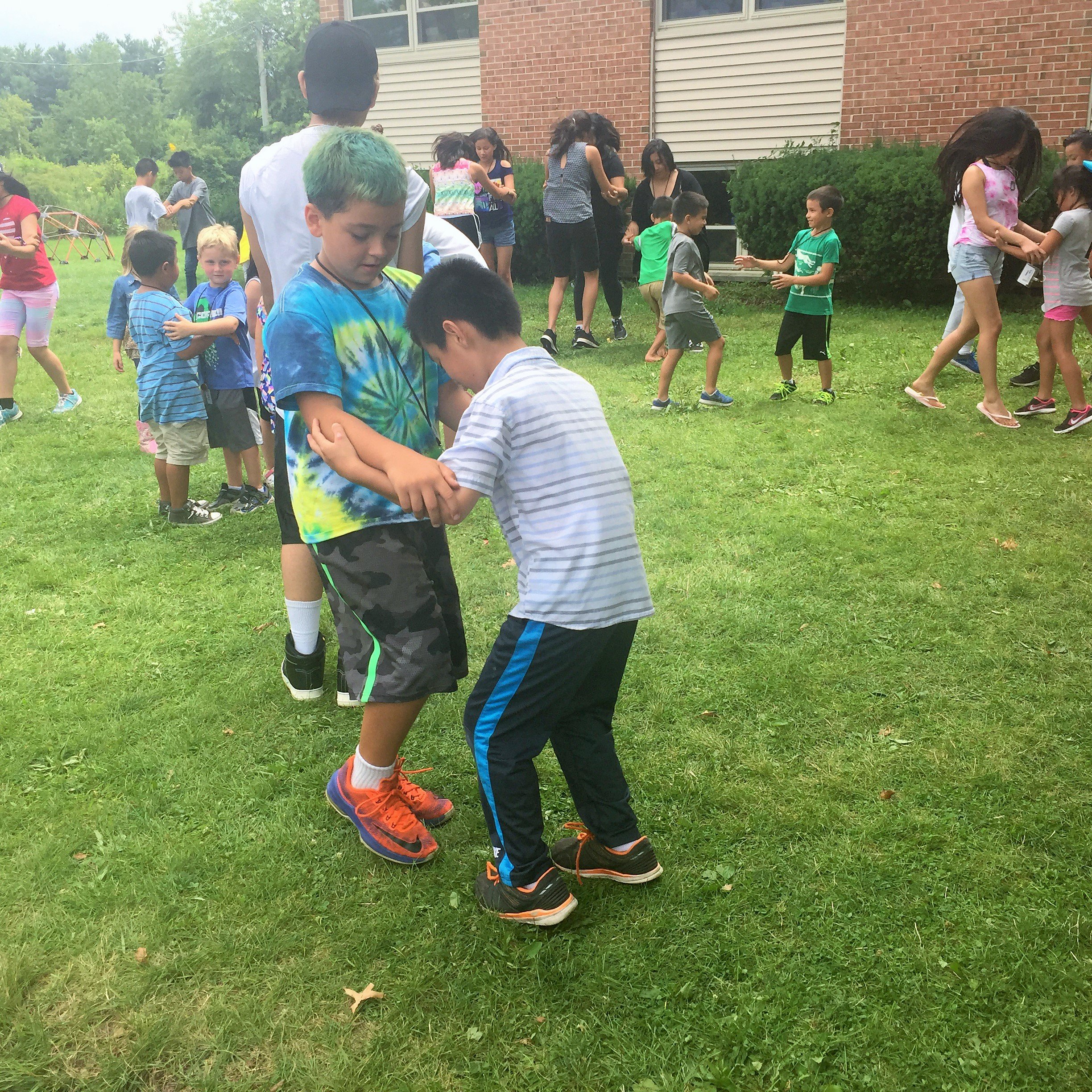 Two boys holding arms and spinning in a game on the grass, with other kids playing behind.