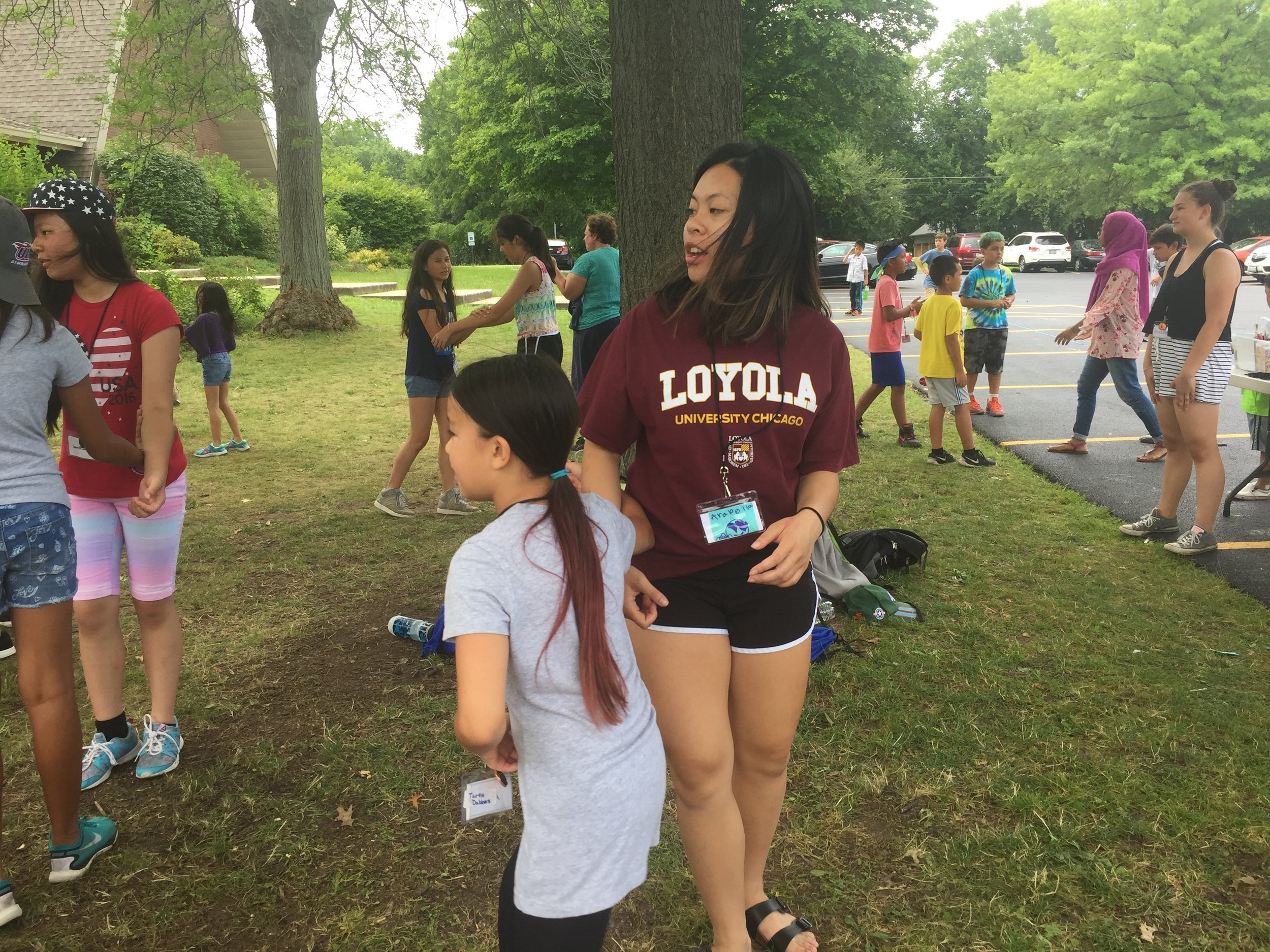 Young woman in a Loyola sweatshirt standing with a girl on a lawn during an outdoor camp day.