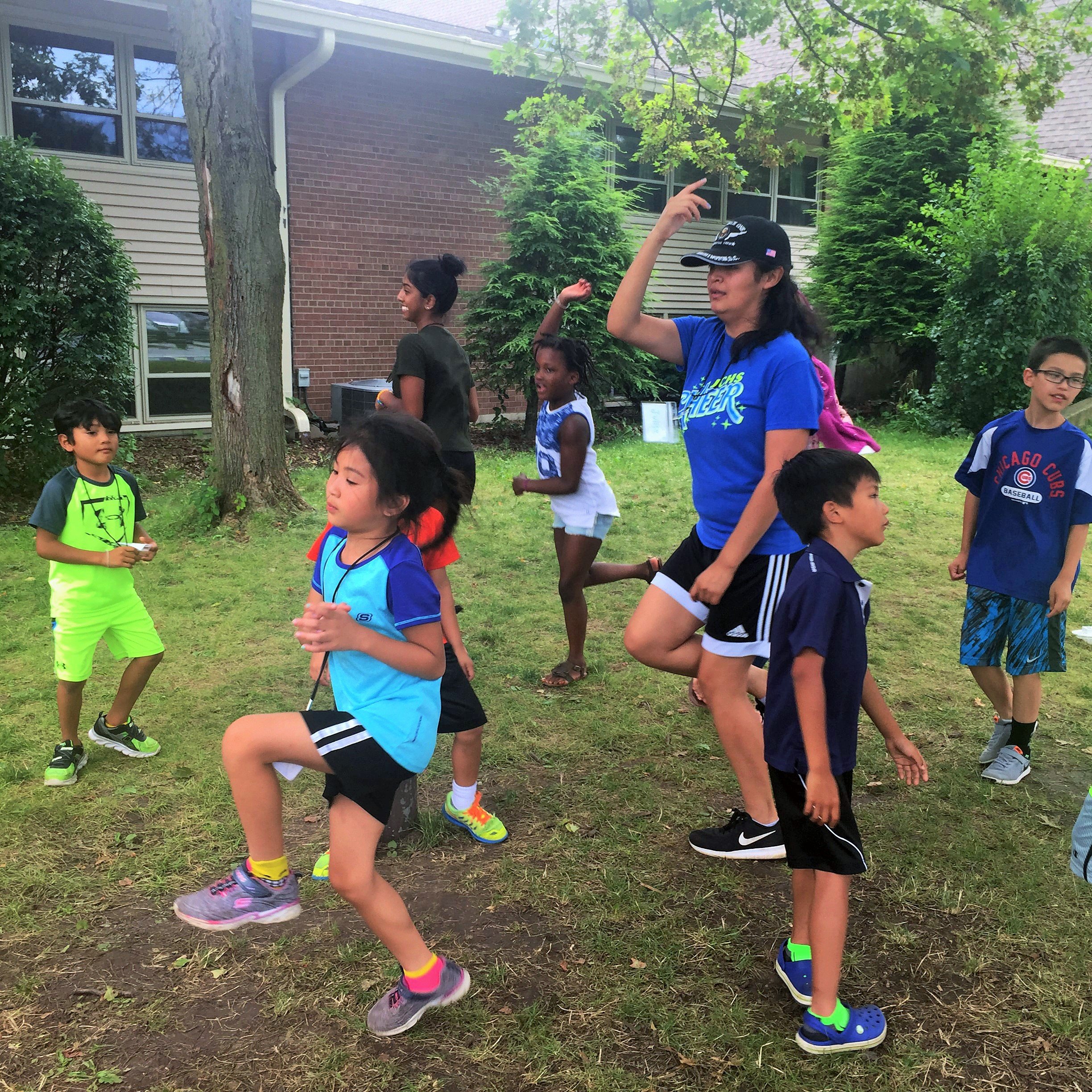 Woman in a cap leading kids in a dance or exercise outdoors, everyone balancing on one leg with an arm raised.