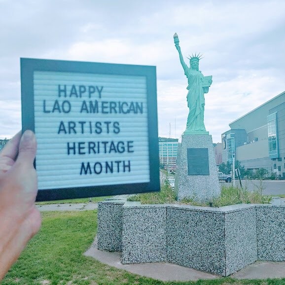 Hand holding a letter board reading Happy Lao American Artists Heritage Month beside a Statue of Liberty replica