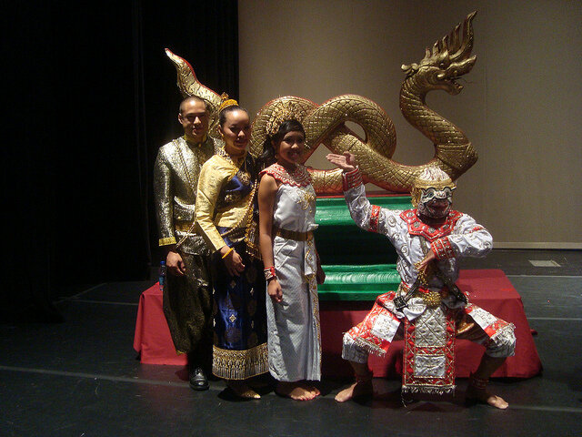 Lam luang performers in ornate Lao costumes, including a masked Hanuman, posing on stage before a golden Nak sculpture